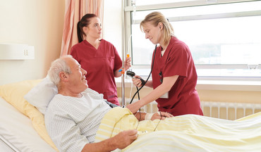 Zwei Pflegerinnen prüfen den Tropf und messen den Blutdruck eines älteren Patienten, welcher im Krankenbett liegt.