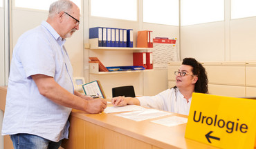 Älterer Patient steht an der Anmeldung und bekommt von der Mitarbeiterin einen Formular zum Ausfüllen ausgehändigt.